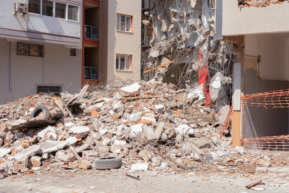 The image depicts a large pile of construction debris and rubble in front of a multi-storey residential building. The rubble consists of broken concrete, bricks, and scattered metal wiring, with various textures and shades of gray, orange, and white. Some debris includes concrete blocks, torn insulation, and bent rebar, indicating demolition activity. In the foreground, two black tires are partially buried among the debris, and the ground appears dusty and uneven. The building behind features several windows with white frames, and one window has a metal security grille. To the right, an orange plastic safety barrier loosely surrounds a section of the debris, suggesting containment for removal. Light and shadows indicate natural daylight, with a clear and unobstructed view of the debris and the urban environment. Rubbish Clearance Tooting provides services to manage such waste, supporting private disposal and on-site clearance of construction debris, which could relate to alternative waste handling solutions outside standard municipal collection methods.