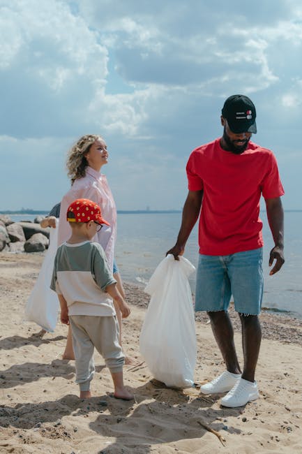 A group of three people, including a man, a woman, and a young boy, are standing on a sandy beach near a body of water, possibly a lake or ocean. The man, dressed in a red T-shirt, ripped denim shorts, white sneakers, and a black cap, is holding a white plastic bag and appears to be collecting litter. The woman, wearing a light pink dress and with curly blonde hair, stands behind them, looking out towards the water. The young boy, dressed in a grey T-shirt, beige shorts, and a red cap with yellow spots, is attentively watching the man. The background shows a partly cloudy sky with some patches of blue, and large rocks lining the shoreline. This scene suggests a voluntary clean-up activity or community effort to remove rubbish from the beach, aligning with private waste handling and alternative rubbish collection services provided by companies like Rubbish Clearance Tooting, who operate in the area to support rubbish removal outside of local authority services.