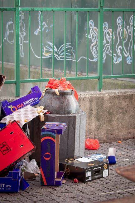 A collection of discarded cardboard boxes and plastic packaging materials, including a purple fruit box and a red crate, is piled around a large metal container situated on a concrete base. The containers are resting on a paved pavement adjacent to a protective green metal fence, behind which a mural with white chalk-like sketches depicting figures and other abstract shapes is visible. Some of the trash, including a red piece of foam and small white plastic items, is scattered on the ground nearby, indicating recent rubbish disposal. The scene appears to be outdoors, possibly in an urban area, with natural daylight illuminating the cluttered area, reflecting a typical scene where independent rubbish clearance services, such as those offered by Rubbish Clearance Tooting, might be called for removing waste from public or private spaces.