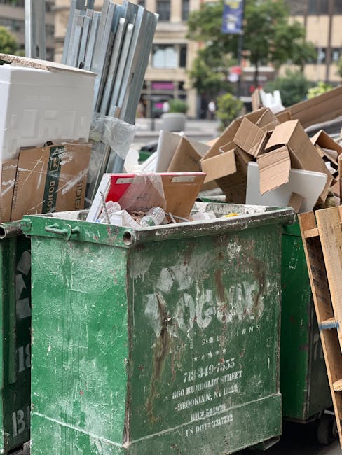 A large, weathered green skip filled with various types of rubbish including cardboard boxes, paper packaging, and miscellaneous waste items, situated outdoors on a paved area. Some cardboard boxes are partially collapsed or torn, and a few plastic bottles and packaging materials are visible among the clutter. The skip is positioned on a street or urban environment, with a background showing blurred trees, buildings, and parked cars, indicating a busy city area. The scene suggests an on-site rubbish clearance operation, possibly carried out by Rubbish Clearance Tooting, for waste removal from flats or commercial premises on High Street. The natural daylight highlights the textures and colors of the materials, emphasizing the varied materials typically associated with private waste handling or alternative rubbish collection services in an urban setting.