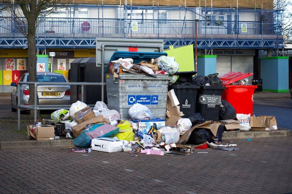 A collection of discarded cardboard boxes and plastic packaging materials, including a purple fruit box and a red crate, is piled around a large metal container situated on a concrete base. The containers are resting on a paved pavement adjacent to a protective green metal fence, behind which a mural with white chalk-like sketches depicting figures and other abstract shapes is visible. Some of the trash, including a red piece of foam and small white plastic items, is scattered on the ground nearby, indicating recent rubbish disposal. The scene appears to be outdoors, possibly in an urban area, with natural daylight illuminating the cluttered area, reflecting a typical scene where independent rubbish clearance services, such as those offered by Rubbish Clearance Tooting, might be called for removing waste from public or private spaces.
